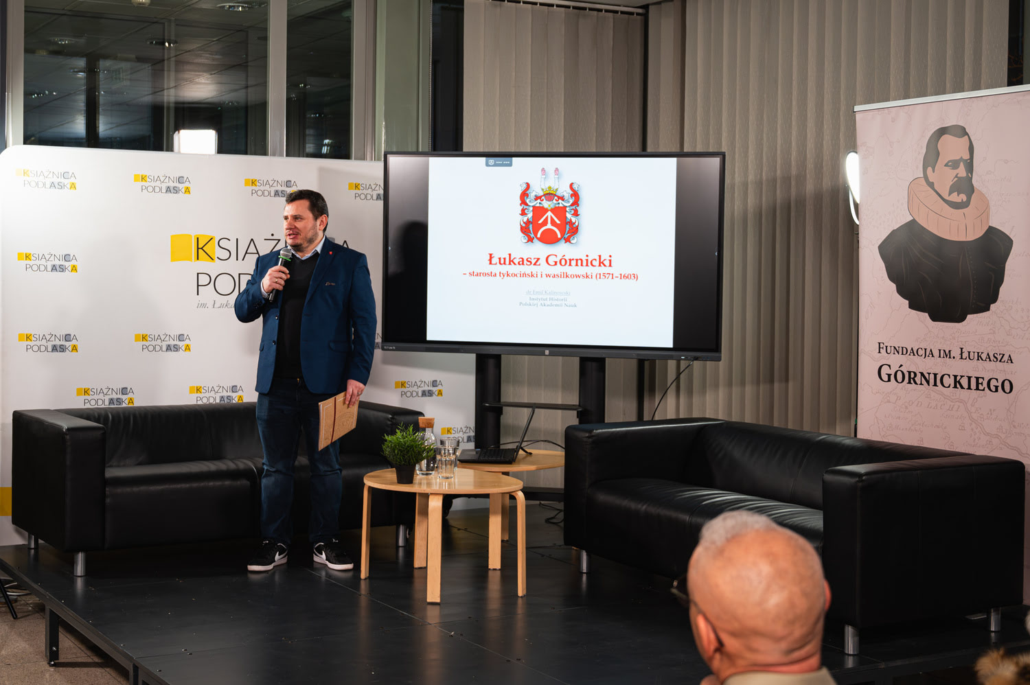 Prelegent stoi na małej scene, za nim roll up Fundacji Górnickiego, ścianka Książnicy Podlaskiej, ekran z prezentacją, dwie sofy oraz stolik.
